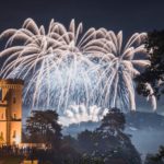 Fireworks light up the night sky behind a castle tower, with spectators gathered to watch the mesmerizing firework display amidst trees and fog.