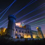 A castle illuminated at night with blue and green laser beams crossing the starry sky above it, enhanced by a spectacular firework display.