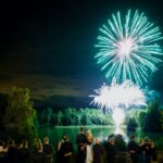 People in formal attire watch a mesmerizing firework display of green and white fireworks illuminate a lake surrounded by trees at night.