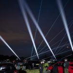 Cars parked outdoors at night with bright, white beams of light illuminating the sky during a spectacular firework display.