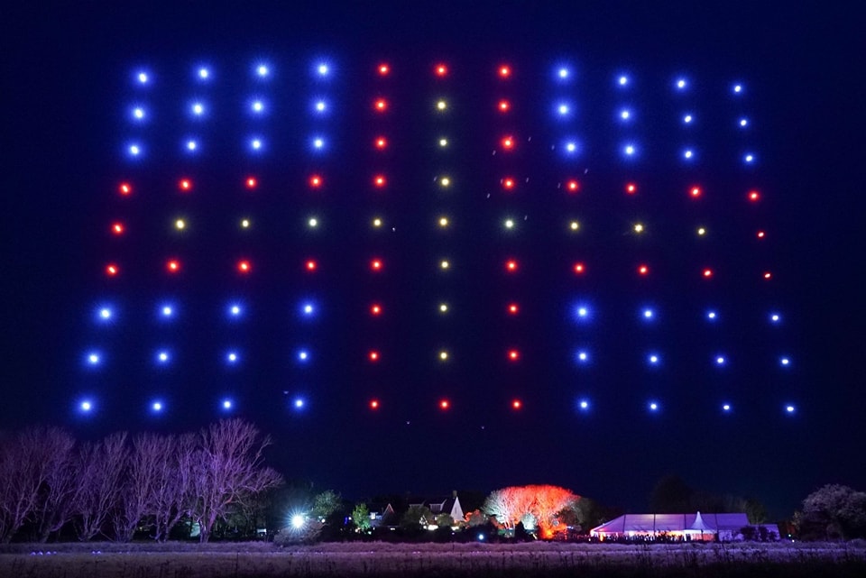 A nighttime display of drones with blue, red, and yellow lights forming a grid pattern over a rural area.