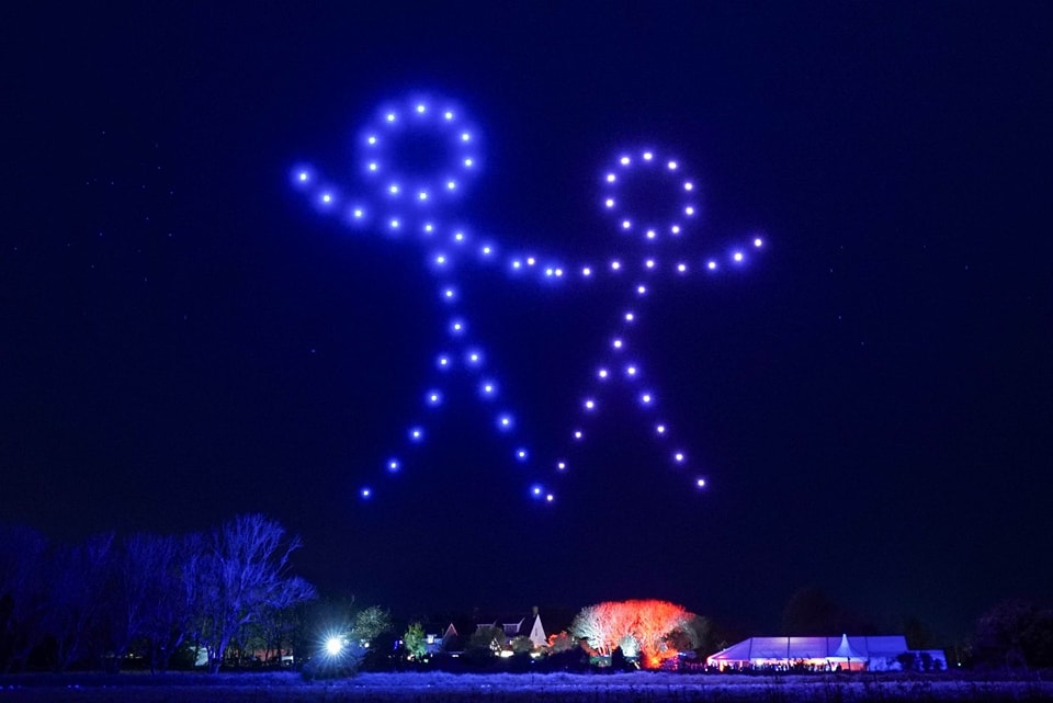 Two figures made of drones light up the night sky, above a landscape with trees and a tent.