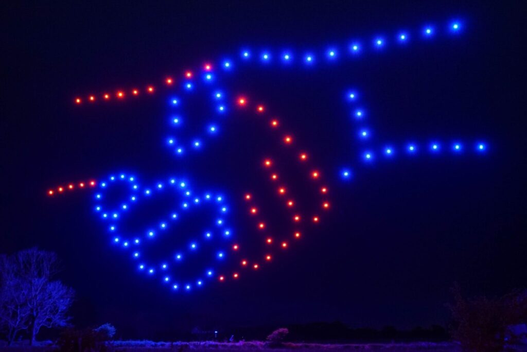 Drones forming an image of a handshake in red and blue lights against a dark sky.