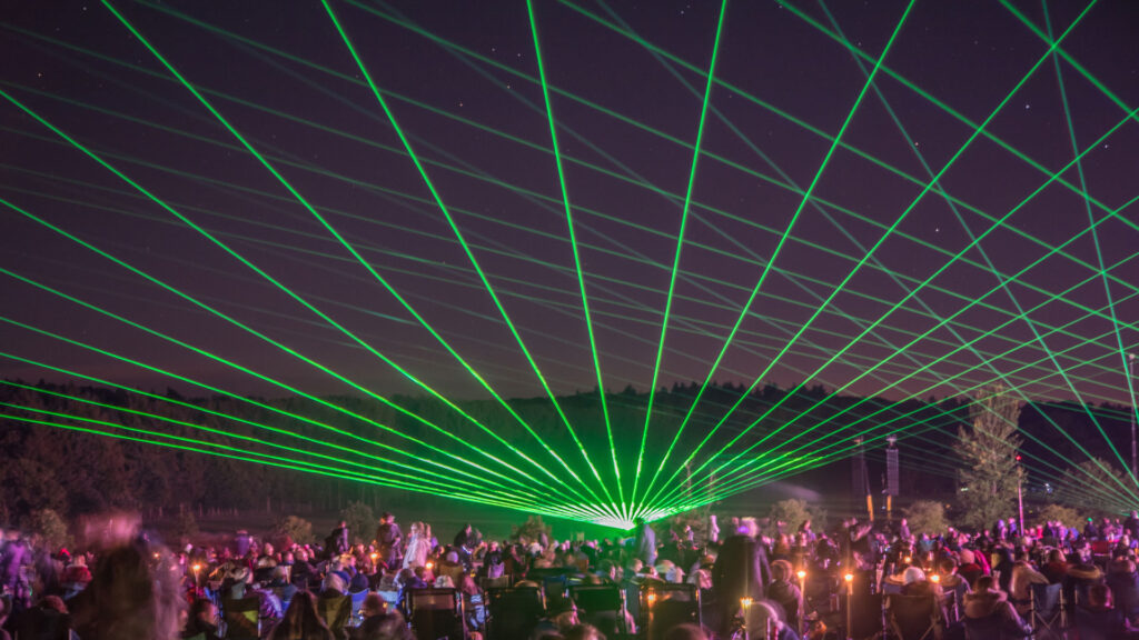 A crowd watches a night sky laser show with green beams under a starry sky.